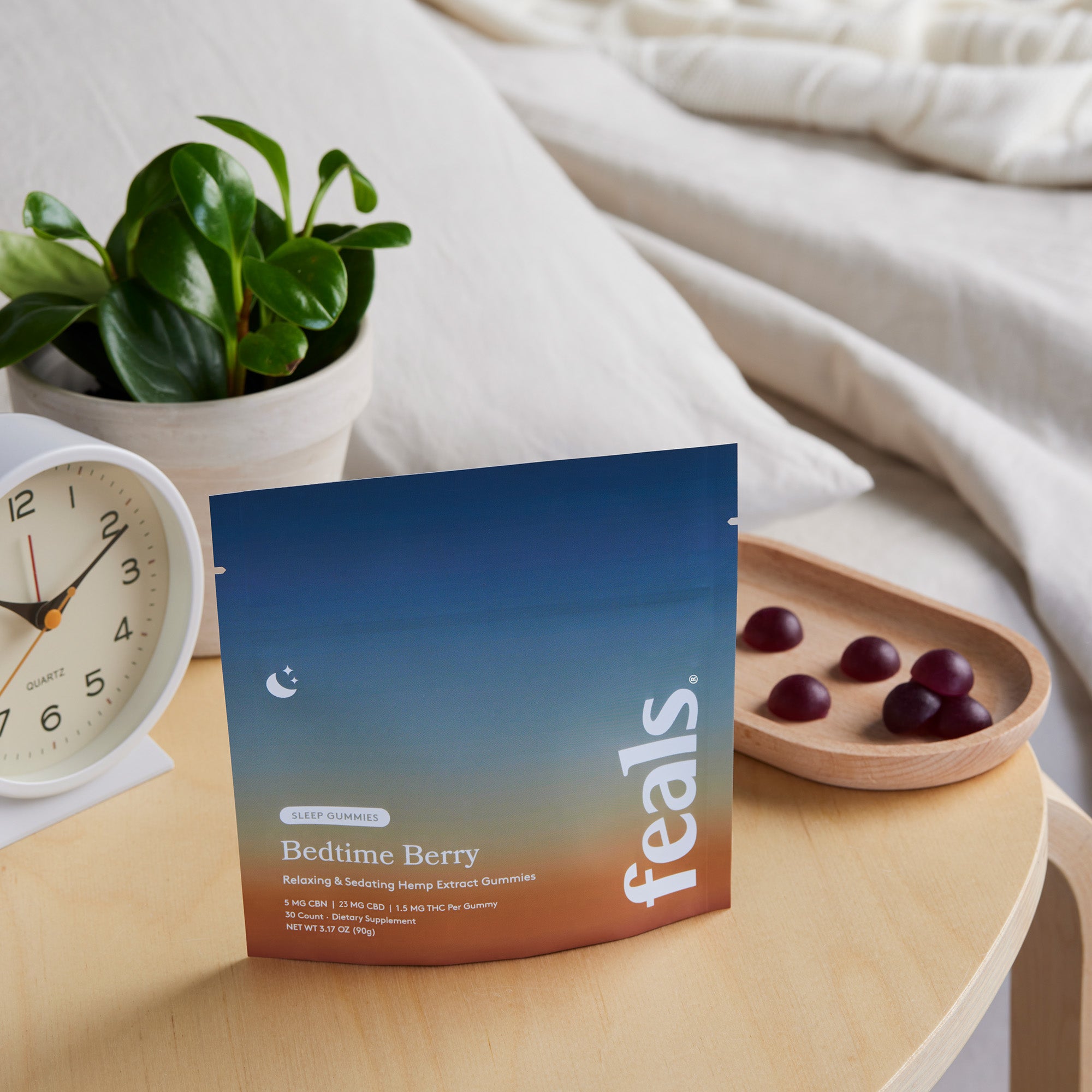 A photo of Feals Bedtime Berry Sleep Gummies on a nightstand with a tray of sleep gummies, an alarm clock, and plant positioned behind it in a bedroom setting.
