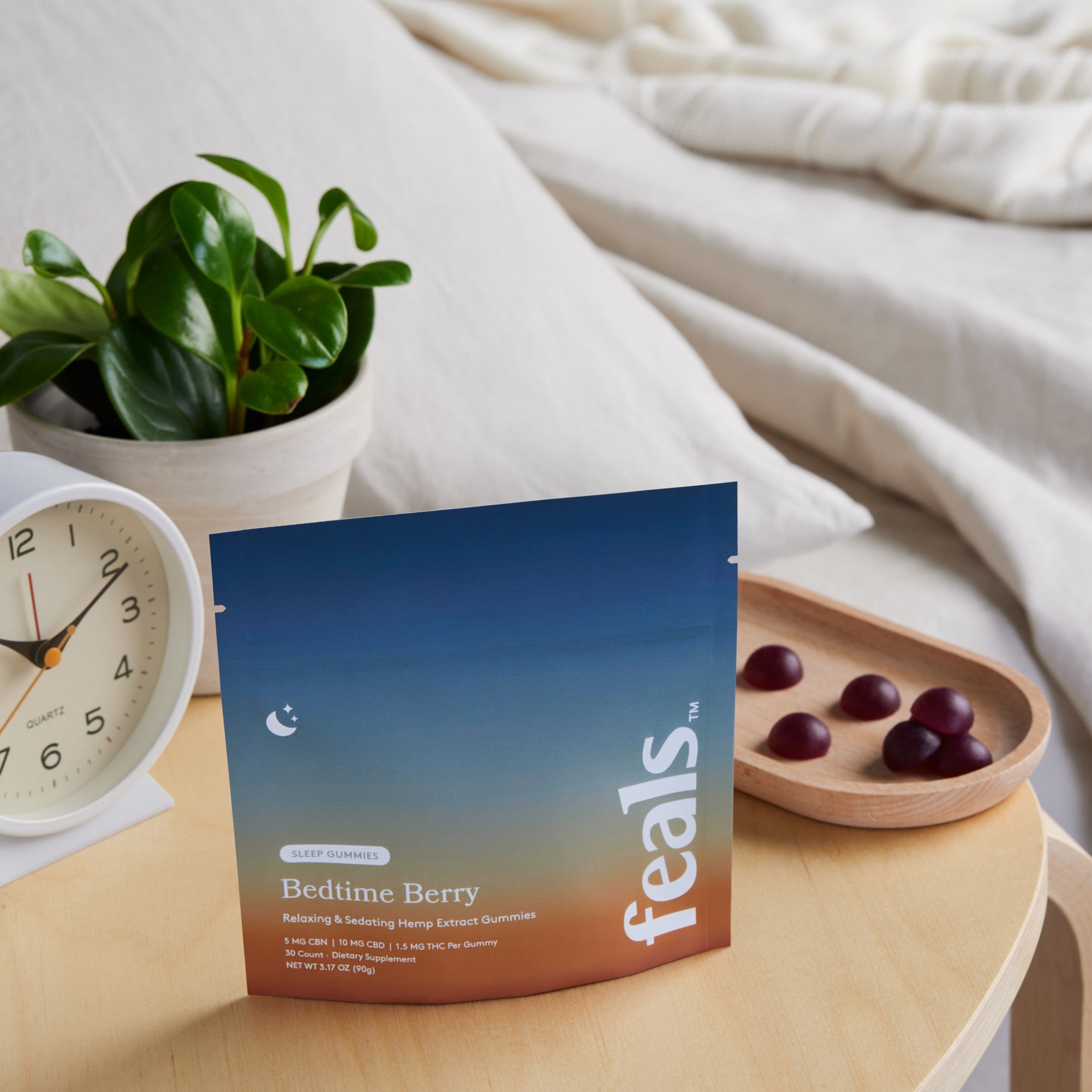 A photo of Feals Bedtime Berry Sleep Gummies on a nightstand with a tray of sleep gummies, an alarm clock, and plant positioned behind it in a bedroom setting.