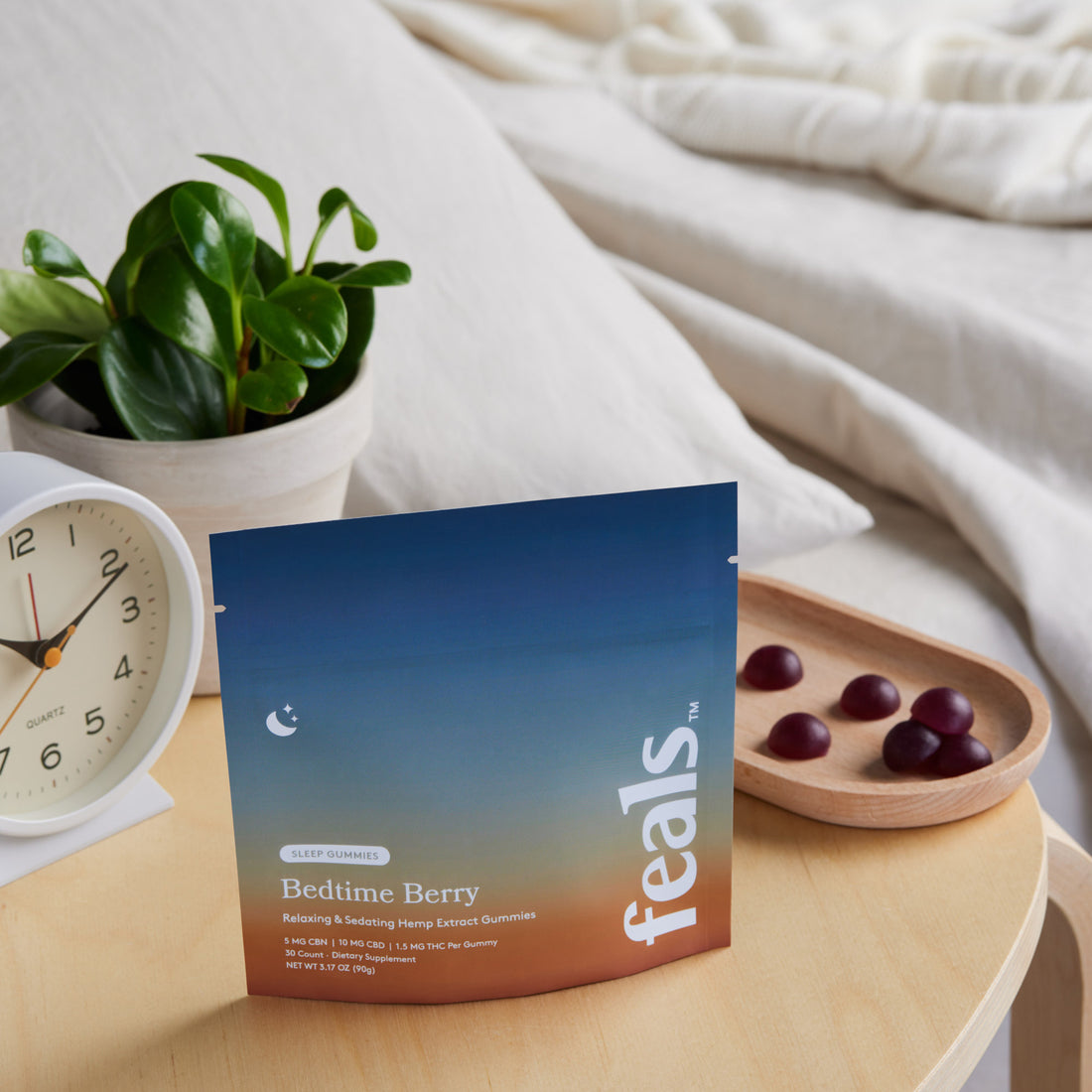 A photo of Feals Bedtime Berry Sleep Gummies on a nightstand with a tray of sleep gummies, an alarm clock, and plant positioned behind it in a bedroom setting.